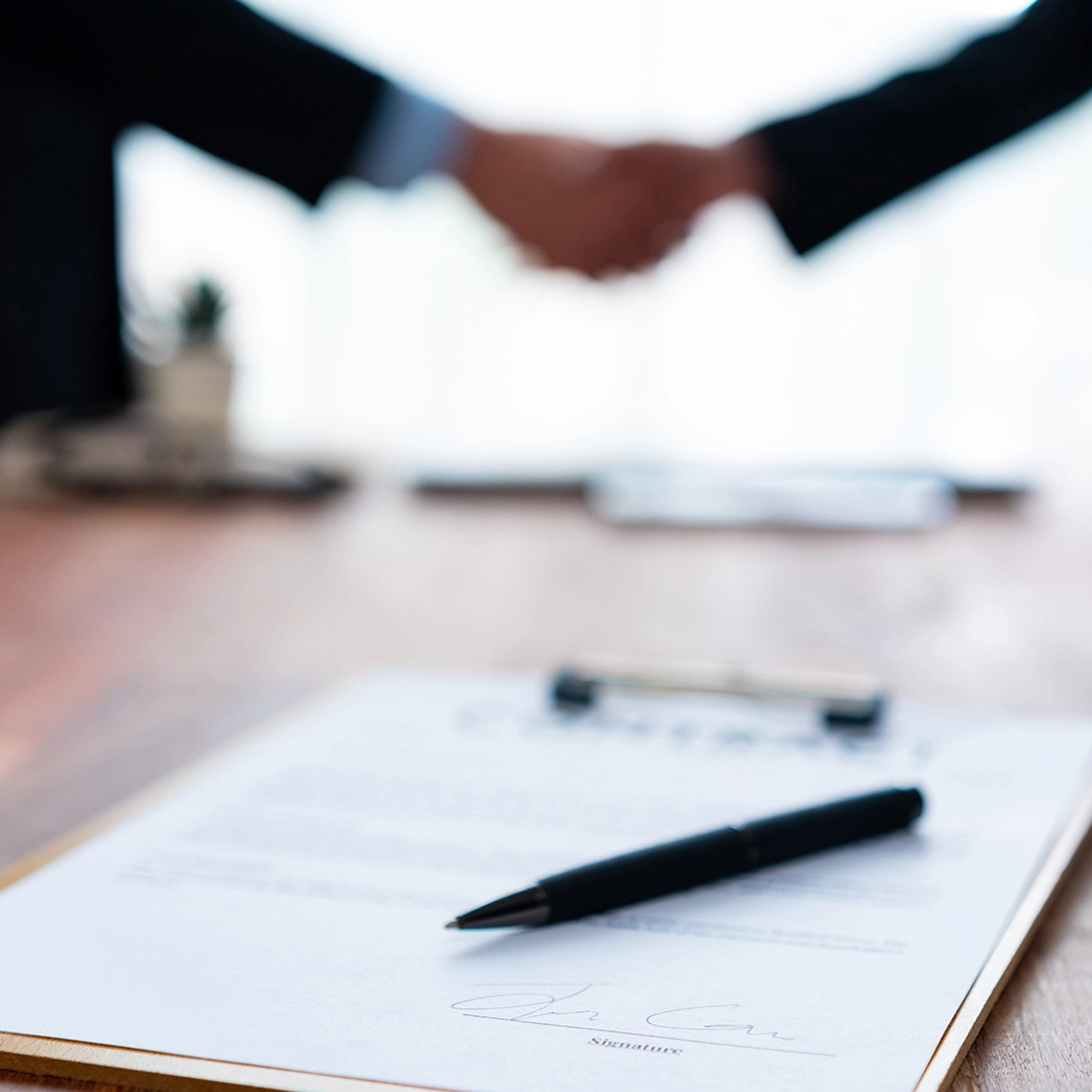 A close-up of a contract with a pen, while two business people shake hands in the background, symbolizing an agreement or partnership. A close-up of a contract with a pen, while two business people shake hands in the background, symbolizing an agreement or partnership.
